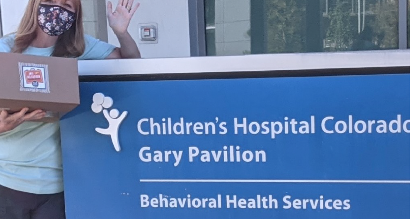 woman holding a box and waving at the camera next to blue Children's Hospital Colorado sign.