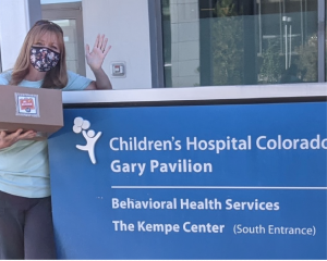 woman holding a box and waving at the camera next to blue Children's Hospital Colorado sign.